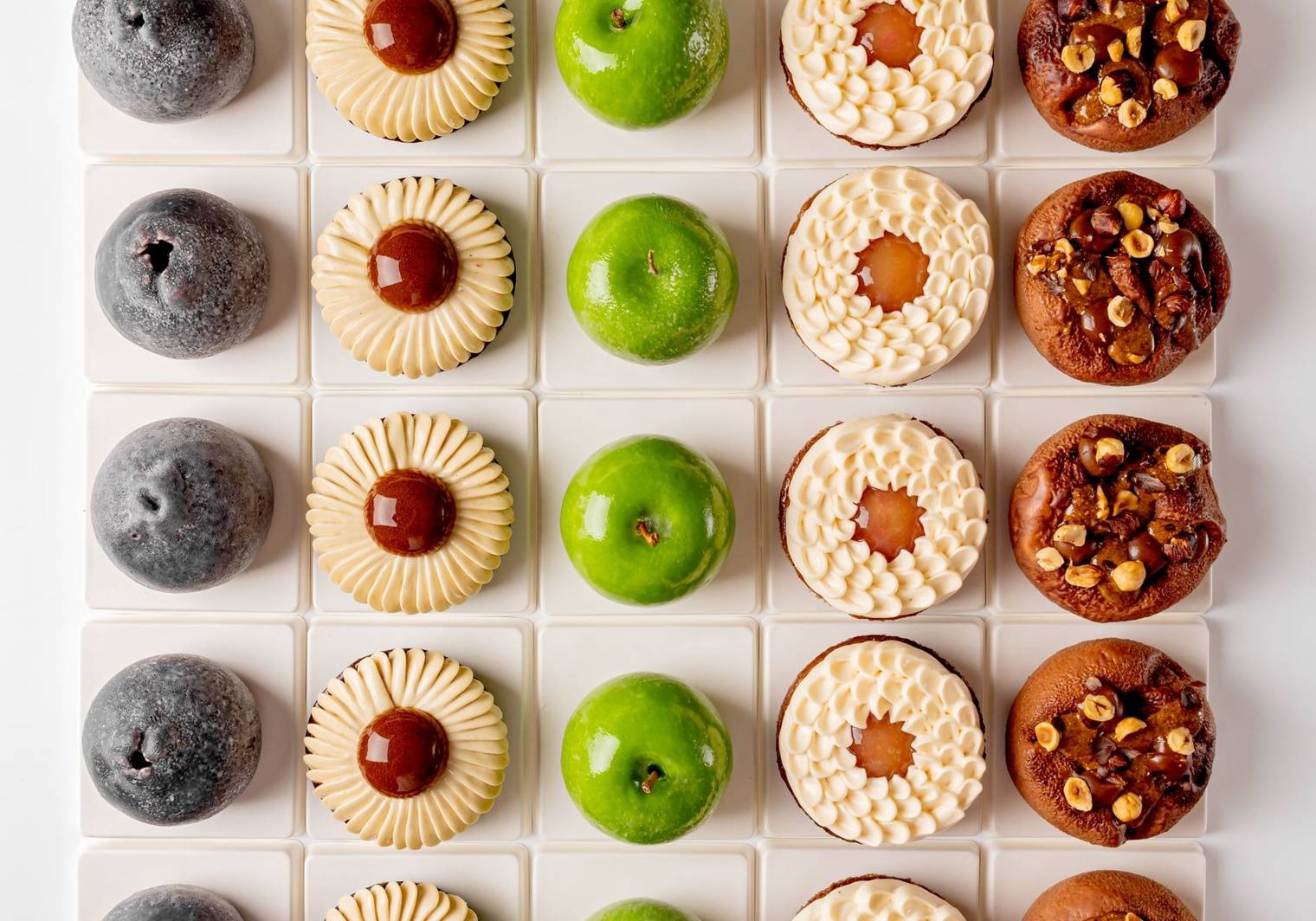 A symmetrical grid of six distinct pastries, showcasing varied textures and colors, from glossy green domes to nut-studded and cream-topped treats, meticulously arranged on white square tiles.