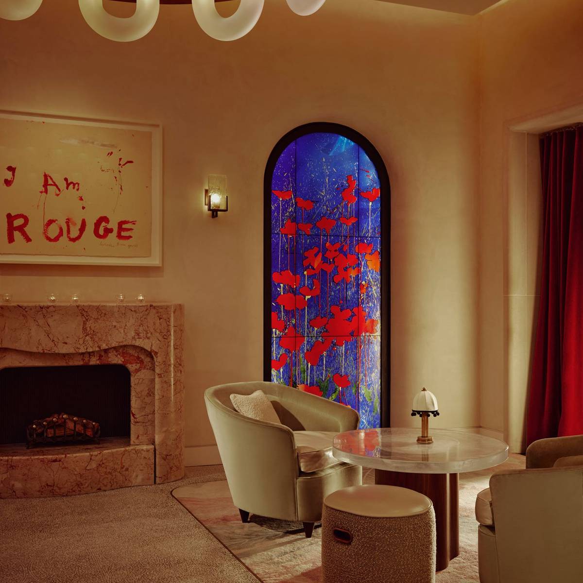 The Connaught Red Room. A round glass table with a small lamp sits to the right. Two beige arm chairs sit either side. In the background there is a blue and red flower art piece. To the left is a marble fireplace