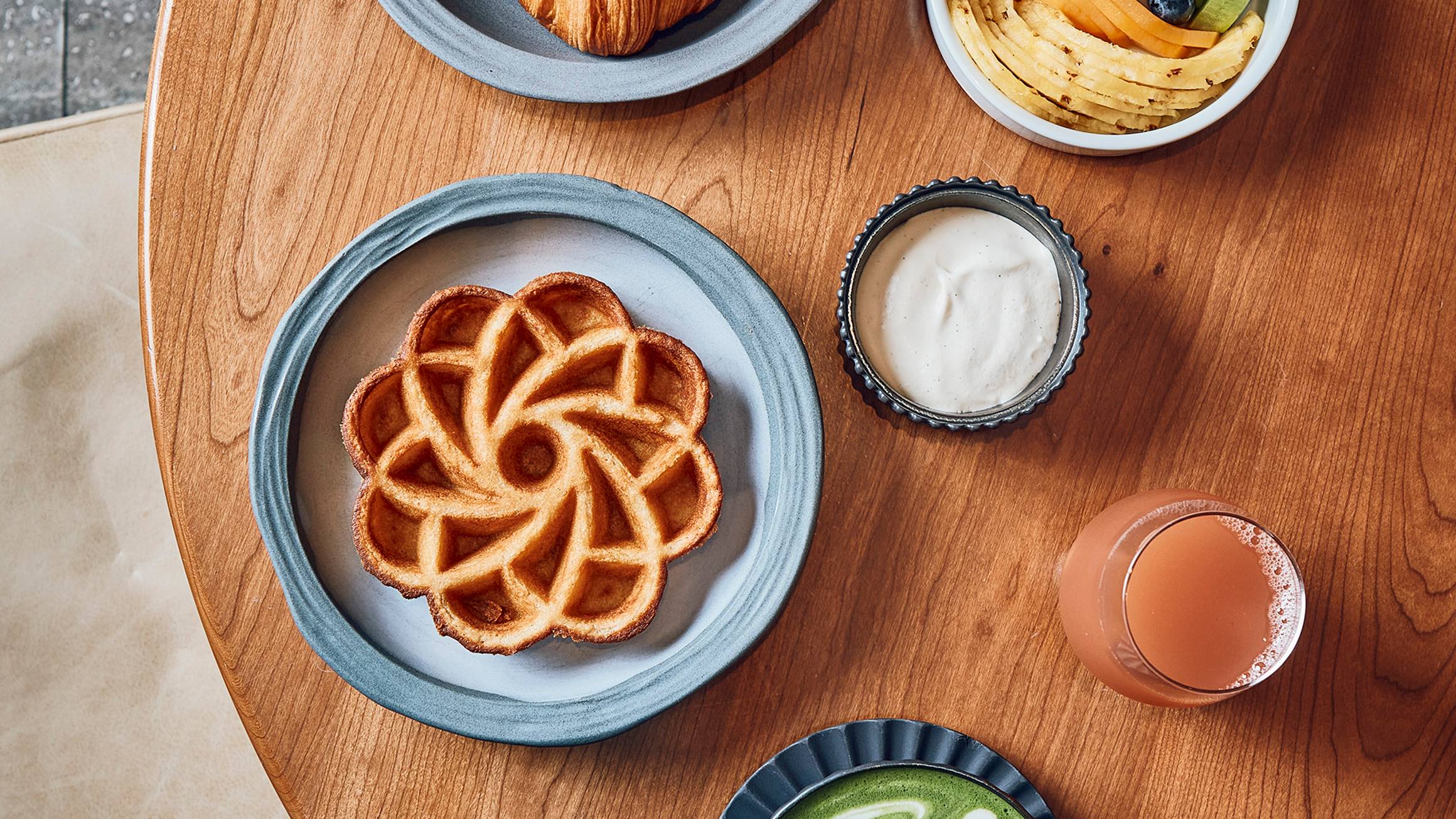 Cedric Grolet breakfast featuring a waffle shaped as a flower, a selection of croissants, a green matcha latte and a bowl of fresh fruit sitting on a wooden table