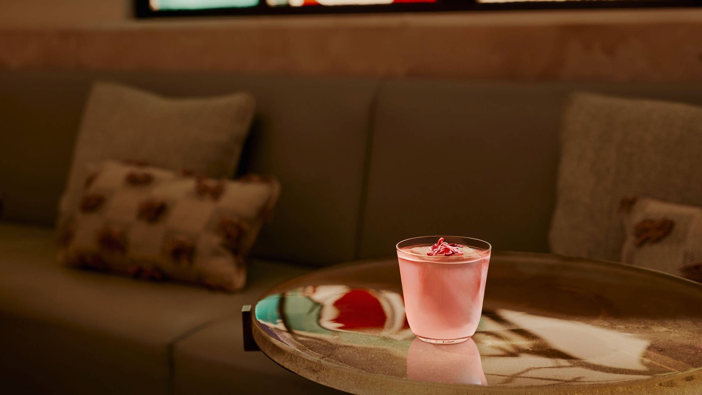 A pink cocktail sits on a glass table. A sofa and cushions are in the background