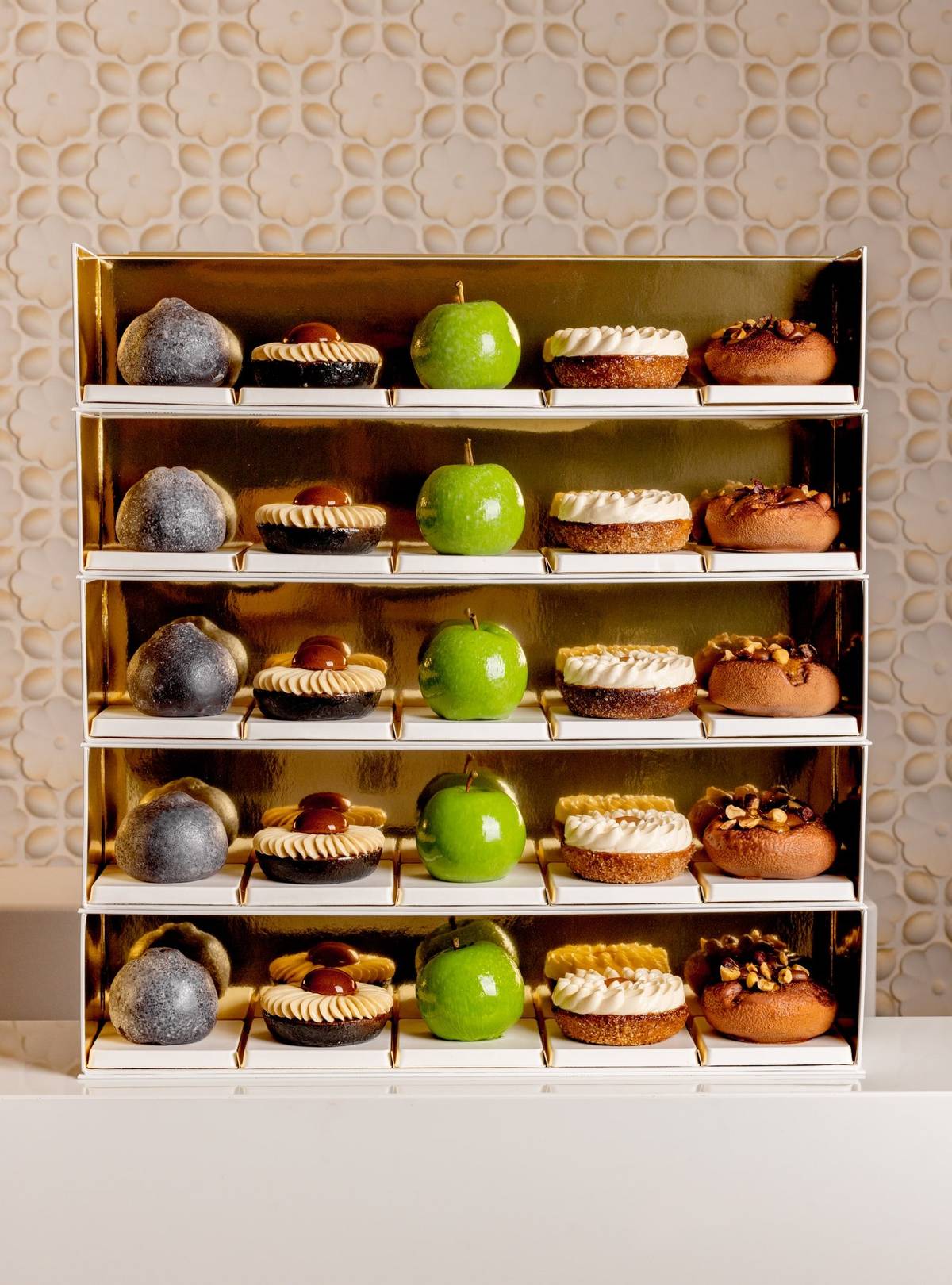 A luxurious display of pastries on golden shelves, featuring green apple-shaped confections, nut-topped treats, and cream-adorned desserts, elegantly arranged against a textured, floral-patterned backdrop.