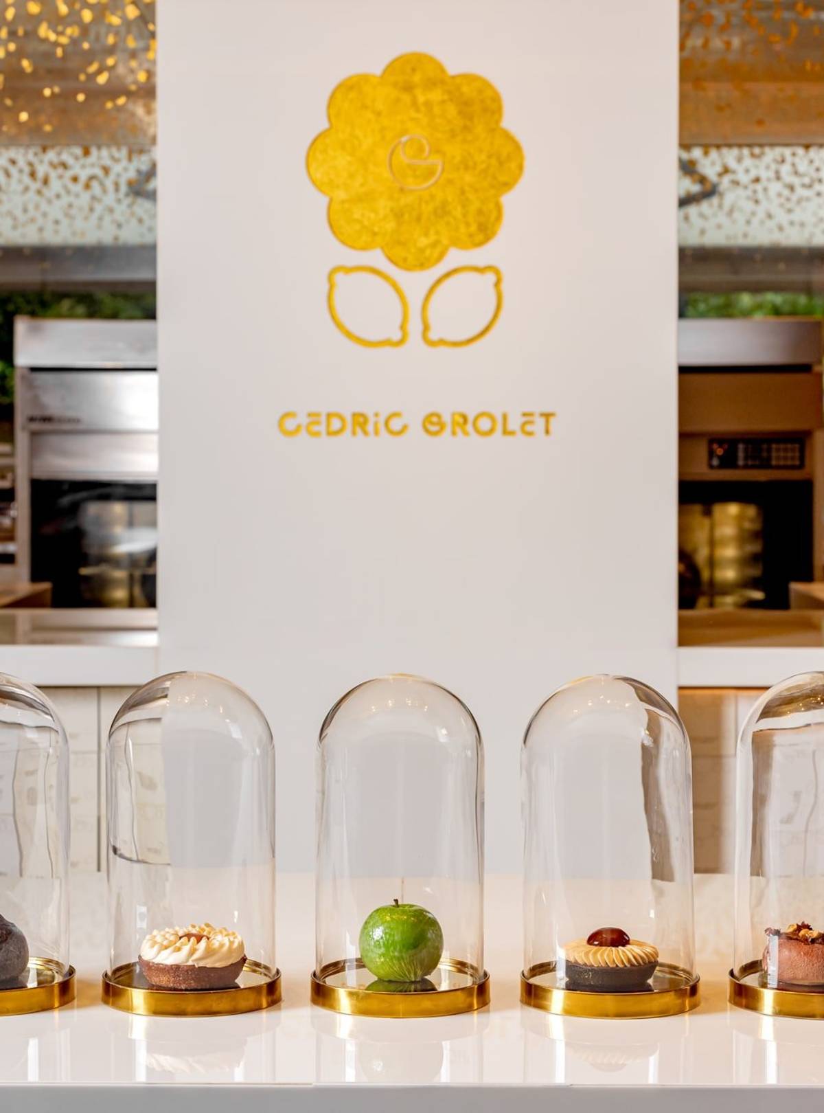 Five exquisite pastries encased in glass domes, elegantly displayed on a minimalist white counter with Cedric Grolet's golden logo in the background.