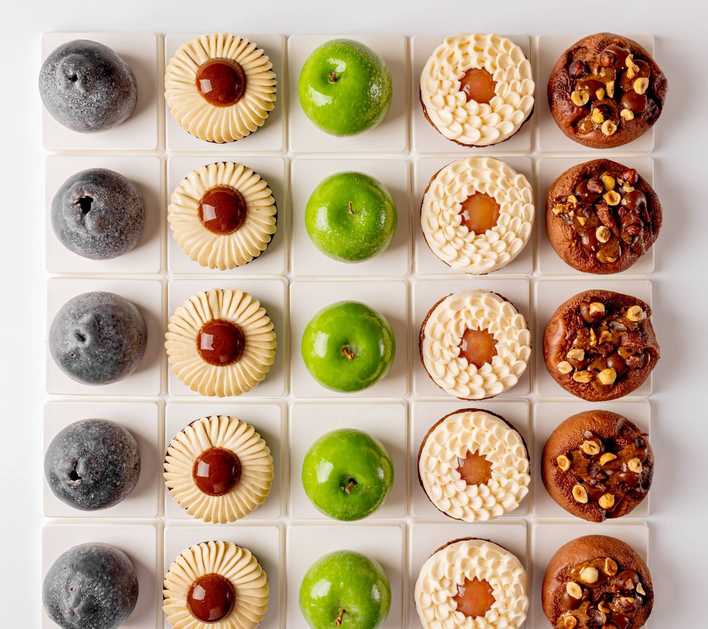 A symmetrical grid of six distinct pastries, showcasing varied textures and colors, from glossy green domes to nut-studded and cream-topped treats, meticulously arranged on white square tiles.