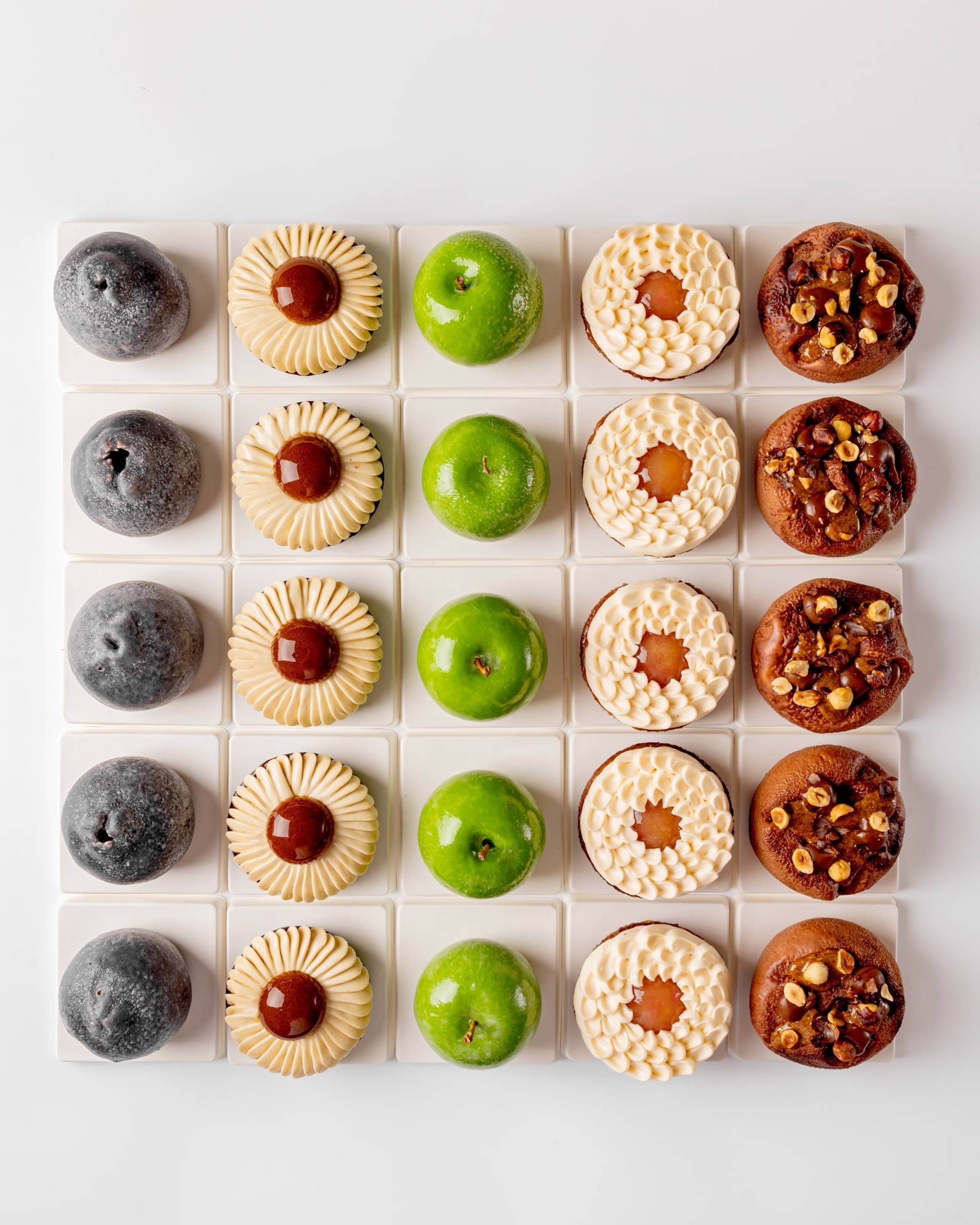 A symmetrical grid of six distinct pastries, showcasing varied textures and colors, from glossy green domes to nut-studded and cream-topped treats, meticulously arranged on white square tiles.