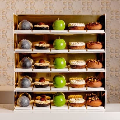 A luxurious display of pastries on golden shelves, featuring green apple-shaped confections, nut-topped treats, and cream-adorned desserts, elegantly arranged against a textured, floral-patterned backdrop.