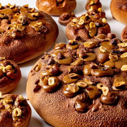 A close-up view of richly textured pastries adorned with caramelized hazelnuts and glossy chocolate accents, creating a warm, indulgent appeal on a smooth white surface.