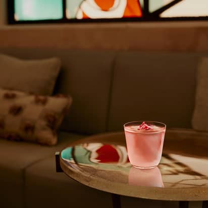 A pink cocktail sits on a glass table. A sofa and cushions are in the background