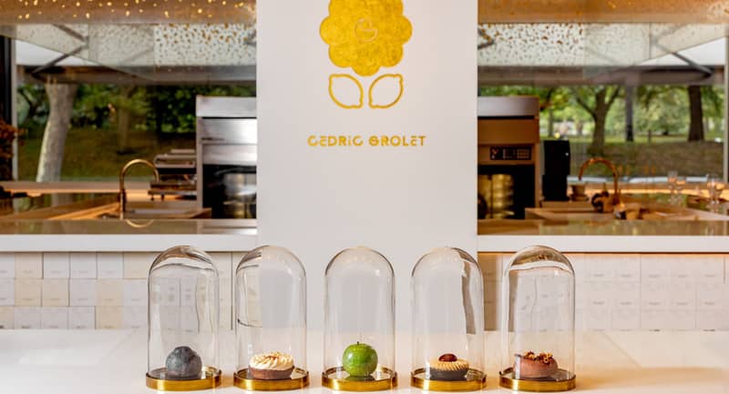 Five exquisite pastries encased in glass domes, elegantly displayed on a minimalist white counter with Cedric Grolet's golden logo in the background.