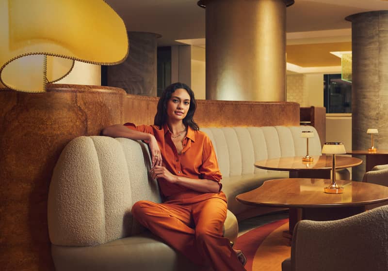 A woman in an elegant orange outfit relaxes on a curved sofa in a warmly lit, stylish lounge setting.