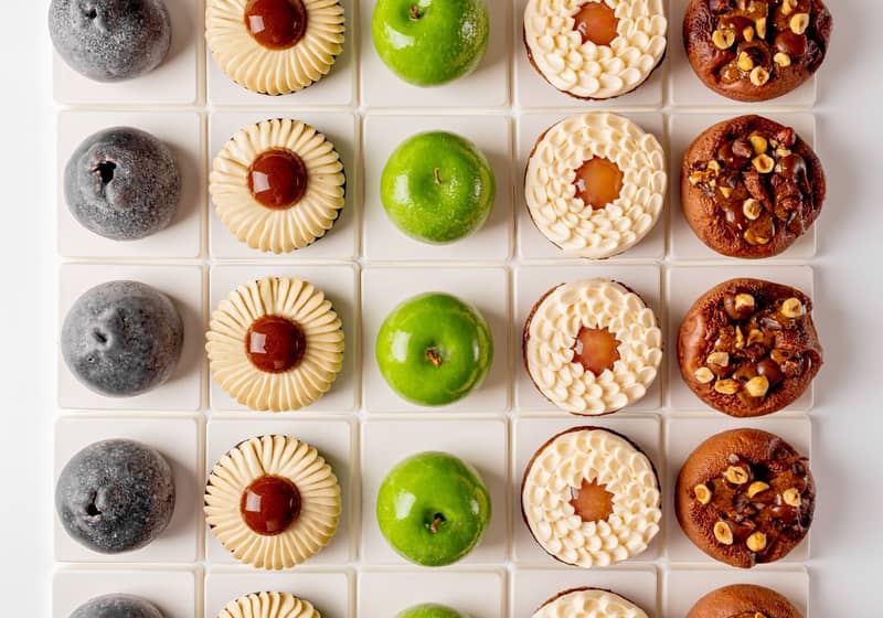 A symmetrical grid of six distinct pastries, showcasing varied textures and colors, from glossy green domes to nut-studded and cream-topped treats, meticulously arranged on white square tiles.