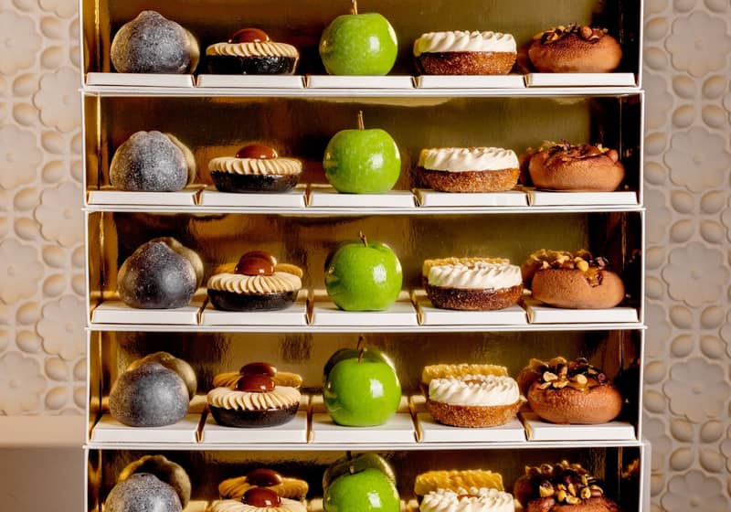 A luxurious display of pastries on golden shelves, featuring green apple-shaped confections, nut-topped treats, and cream-adorned desserts, elegantly arranged against a textured, floral-patterned backdrop.