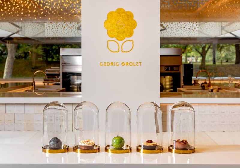 Five exquisite pastries encased in glass domes, elegantly displayed on a minimalist white counter with Cedric Grolet's golden logo in the background.