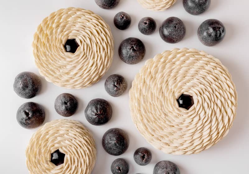 An artistic arrangement of intricately layered pastries with spiral patterns, paired with dark, round fruit-like elements on a pristine white background, creating a visually striking contrast.