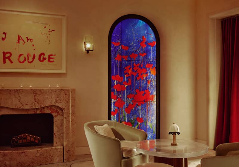 The Connaught Red Room. A round glass table with a small lamp sits to the right. Two beige arm chairs sit either side. In the background there is a blue and red flower art piece. To the left is a marble fireplace