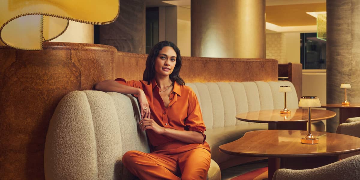 A woman in an elegant orange outfit relaxes on a curved sofa in a warmly lit, stylish lounge setting.