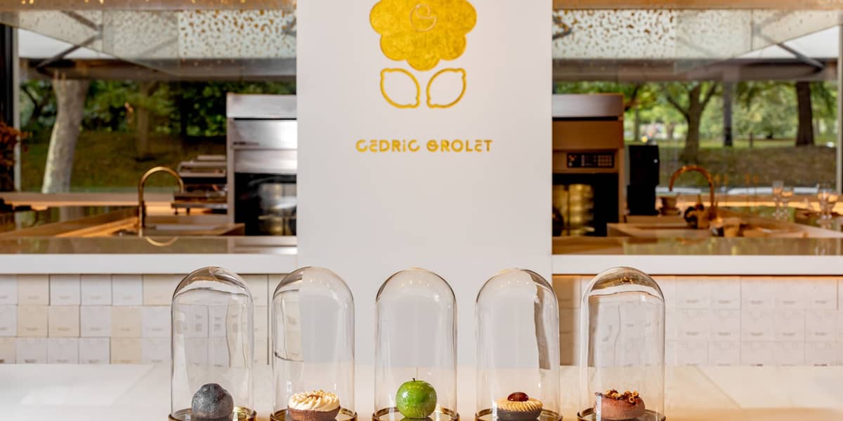 Five exquisite pastries encased in glass domes, elegantly displayed on a minimalist white counter with Cedric Grolet's golden logo in the background.