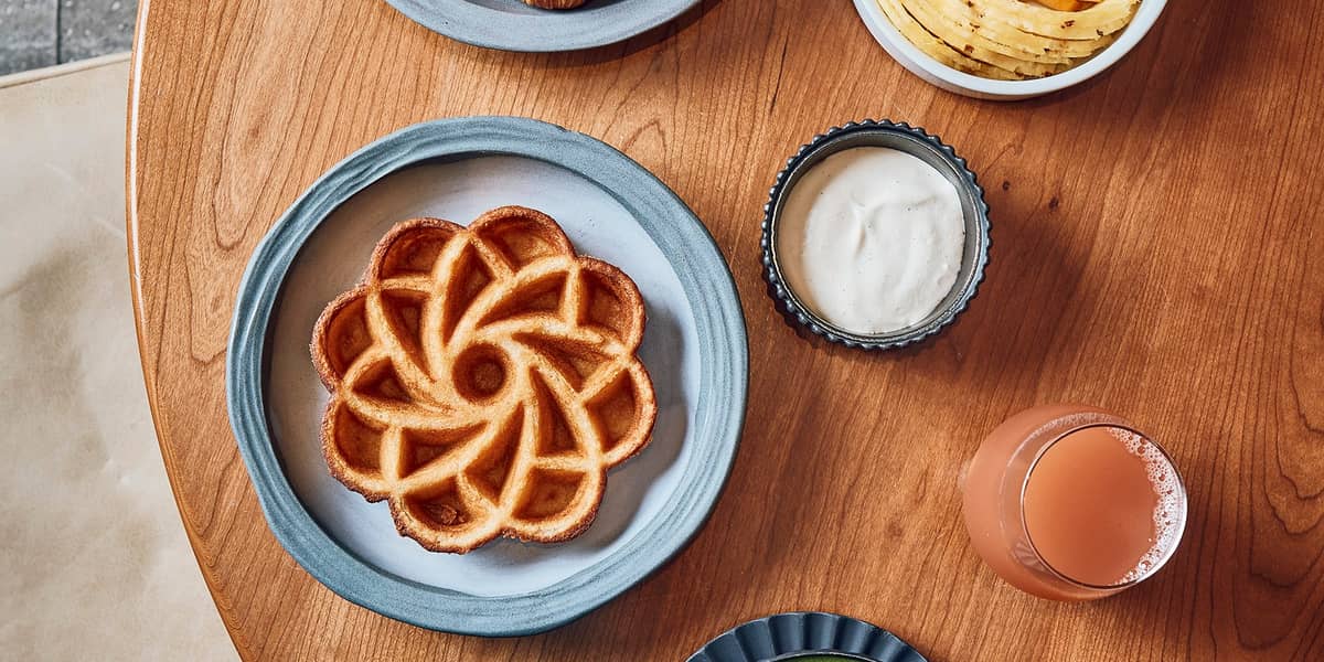 Cedric Grolet breakfast featuring a waffle shaped as a flower, a selection of croissants, a green matcha latte and a bowl of fresh fruit sitting on a wooden table