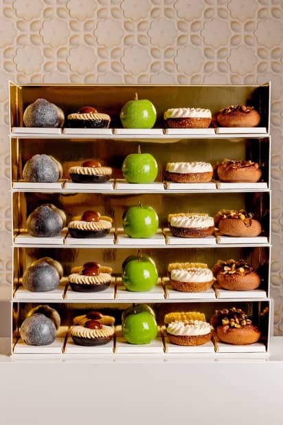 A luxurious display of pastries on golden shelves, featuring green apple-shaped confections, nut-topped treats, and cream-adorned desserts, elegantly arranged against a textured, floral-patterned backdrop.