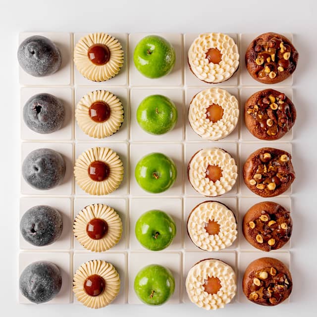 A symmetrical grid of six distinct pastries, showcasing varied textures and colors, from glossy green domes to nut-studded and cream-topped treats, meticulously arranged on white square tiles.