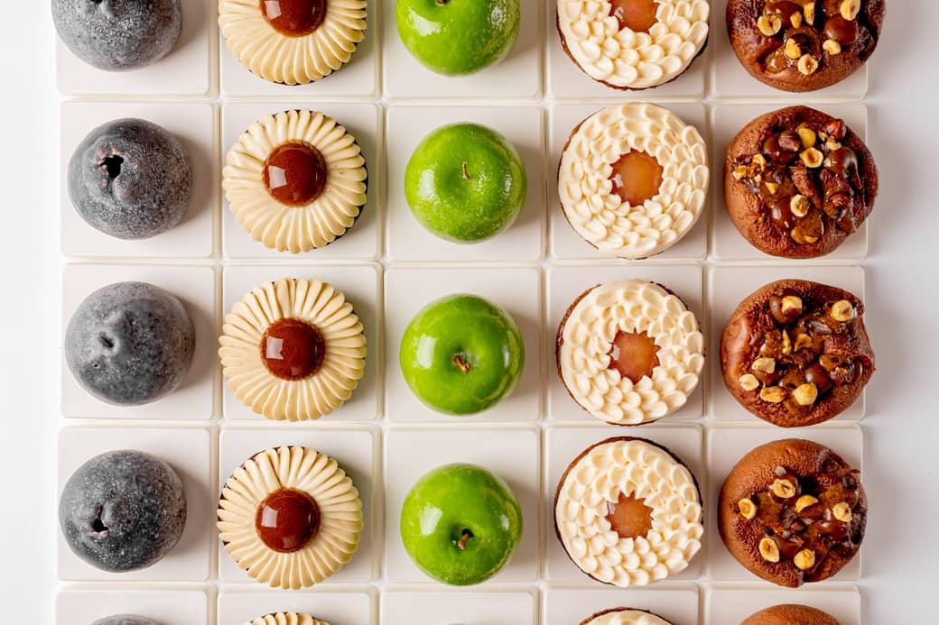 A symmetrical grid of six distinct pastries, showcasing varied textures and colors, from glossy green domes to nut-studded and cream-topped treats, meticulously arranged on white square tiles.