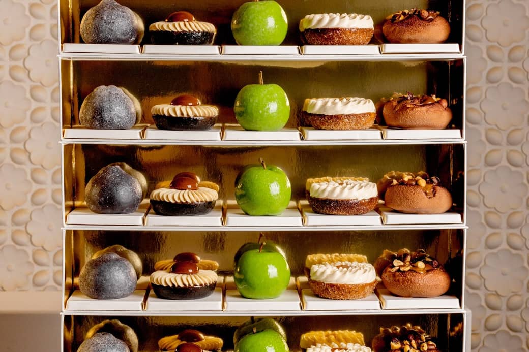 A luxurious display of pastries on golden shelves, featuring green apple-shaped confections, nut-topped treats, and cream-adorned desserts, elegantly arranged against a textured, floral-patterned backdrop.