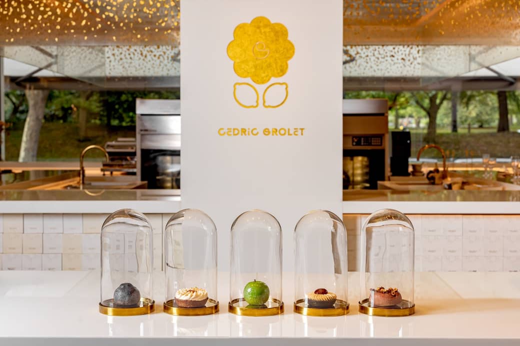 Five exquisite pastries encased in glass domes, elegantly displayed on a minimalist white counter with Cedric Grolet's golden logo in the background.