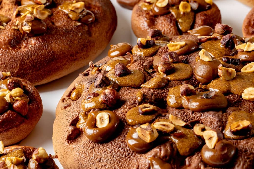 A close-up view of richly textured pastries adorned with caramelized hazelnuts and glossy chocolate accents, creating a warm, indulgent appeal on a smooth white surface.