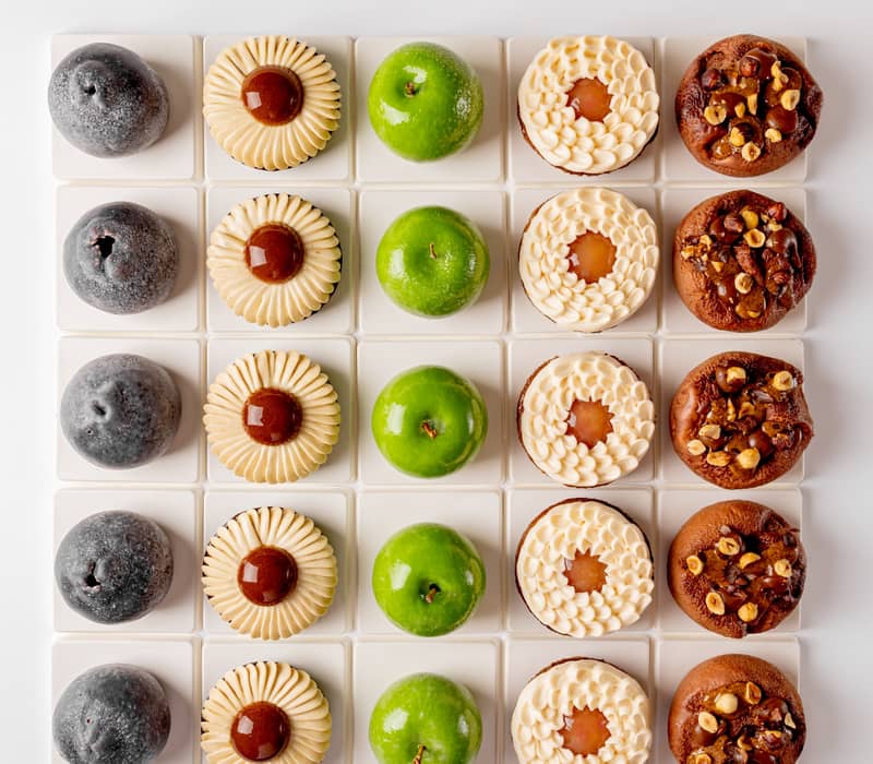 A symmetrical grid of six distinct pastries, showcasing varied textures and colors, from glossy green domes to nut-studded and cream-topped treats, meticulously arranged on white square tiles.