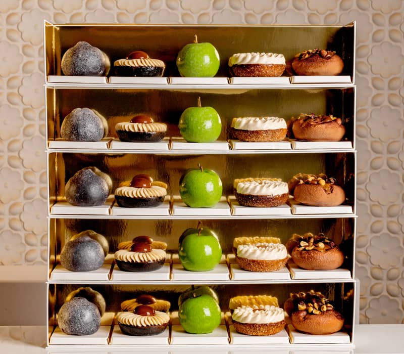 A luxurious display of pastries on golden shelves, featuring green apple-shaped confections, nut-topped treats, and cream-adorned desserts, elegantly arranged against a textured, floral-patterned backdrop.