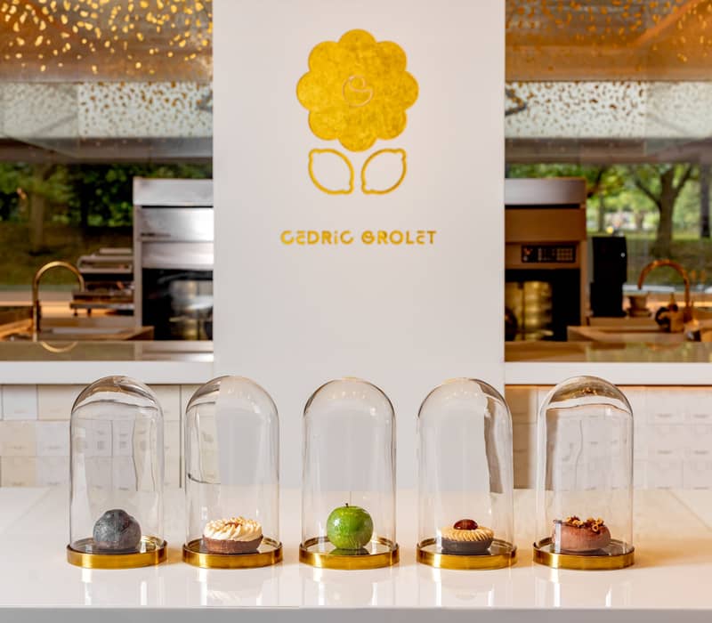 Five exquisite pastries encased in glass domes, elegantly displayed on a minimalist white counter with Cedric Grolet's golden logo in the background.