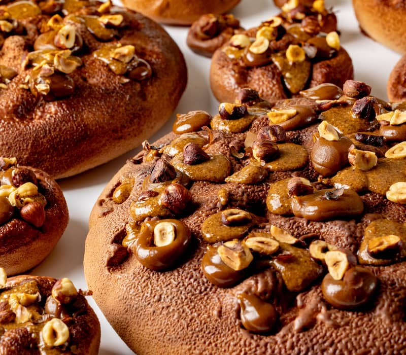 A close-up view of richly textured pastries adorned with caramelized hazelnuts and glossy chocolate accents, creating a warm, indulgent appeal on a smooth white surface.