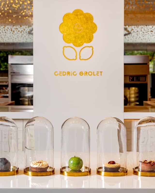 Five exquisite pastries encased in glass domes, elegantly displayed on a minimalist white counter with Cedric Grolet's golden logo in the background.