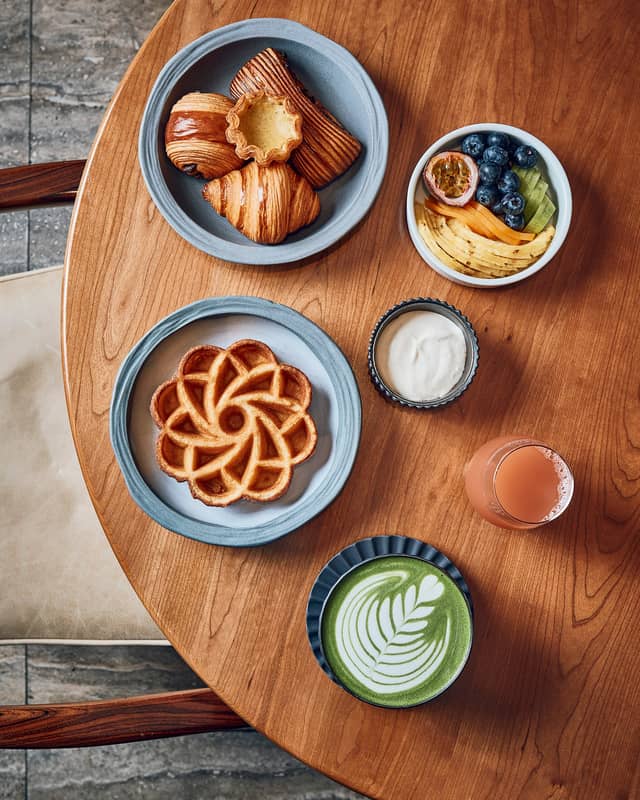 Cedric Grolet breakfast featuring a waffle shaped as a flower, a selection of croissants, a green matcha latte and a bowl of fresh fruit sitting on a wooden table