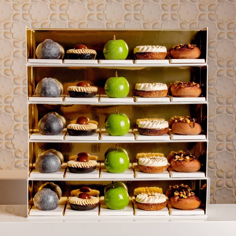 A luxurious display of pastries on golden shelves, featuring green apple-shaped confections, nut-topped treats, and cream-adorned desserts, elegantly arranged against a textured, floral-patterned backdrop.