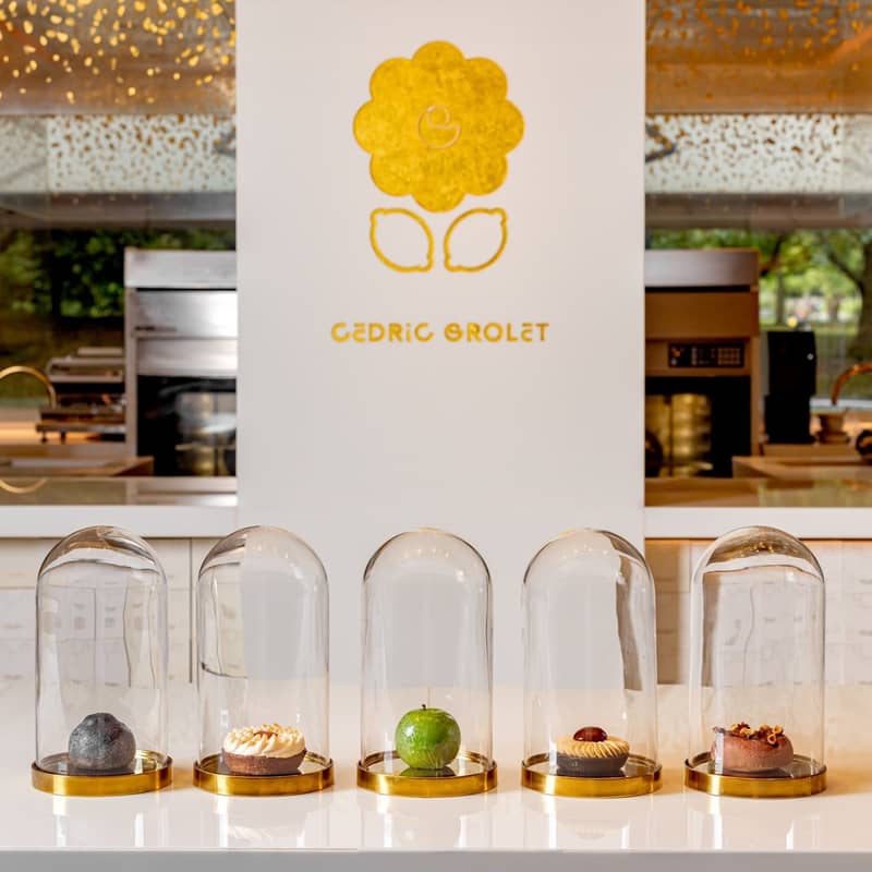 Five exquisite pastries encased in glass domes, elegantly displayed on a minimalist white counter with Cedric Grolet's golden logo in the background.