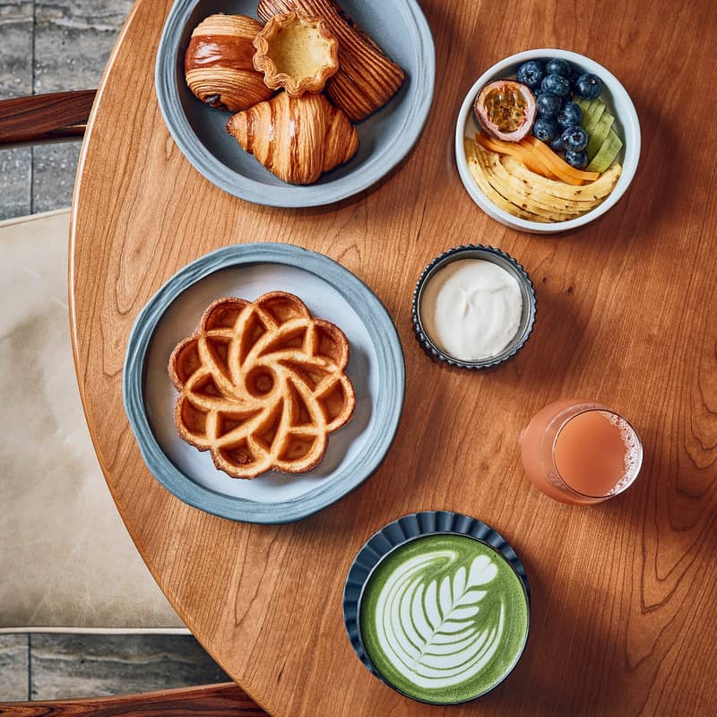 Cedric Grolet breakfast featuring a waffle shaped as a flower, a selection of croissants, a green matcha latte and a bowl of fresh fruit sitting on a wooden table