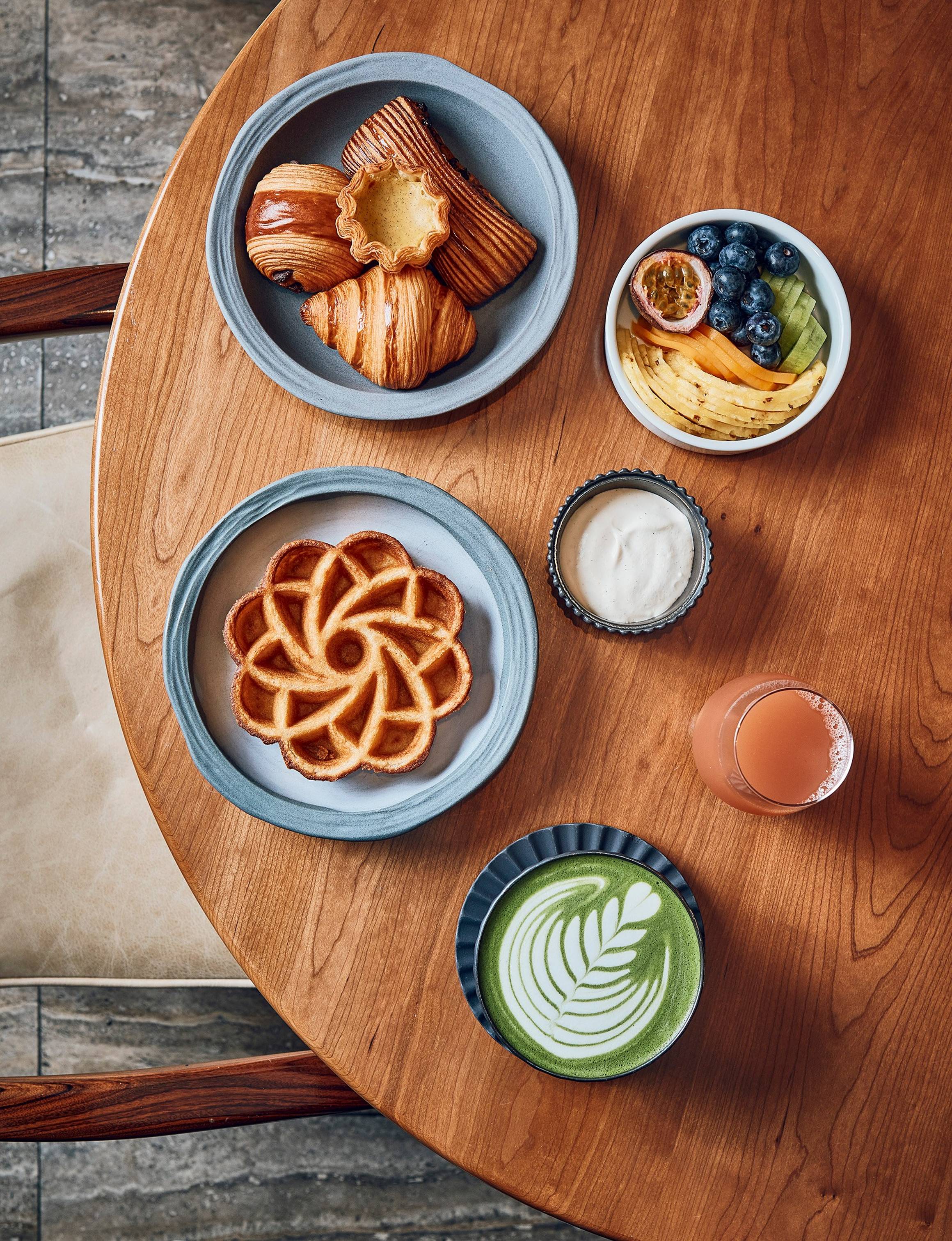 Cedric Grolet breakfast featuring a waffle shaped as a flower, a selection of croissants, a green matcha latte and a bowl of fresh fruit sitting on a wooden table