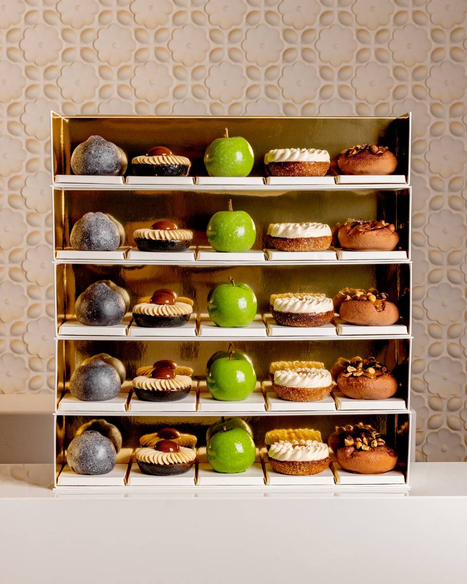 A luxurious display of pastries on golden shelves, featuring green apple-shaped confections, nut-topped treats, and cream-adorned desserts, elegantly arranged against a textured, floral-patterned backdrop.