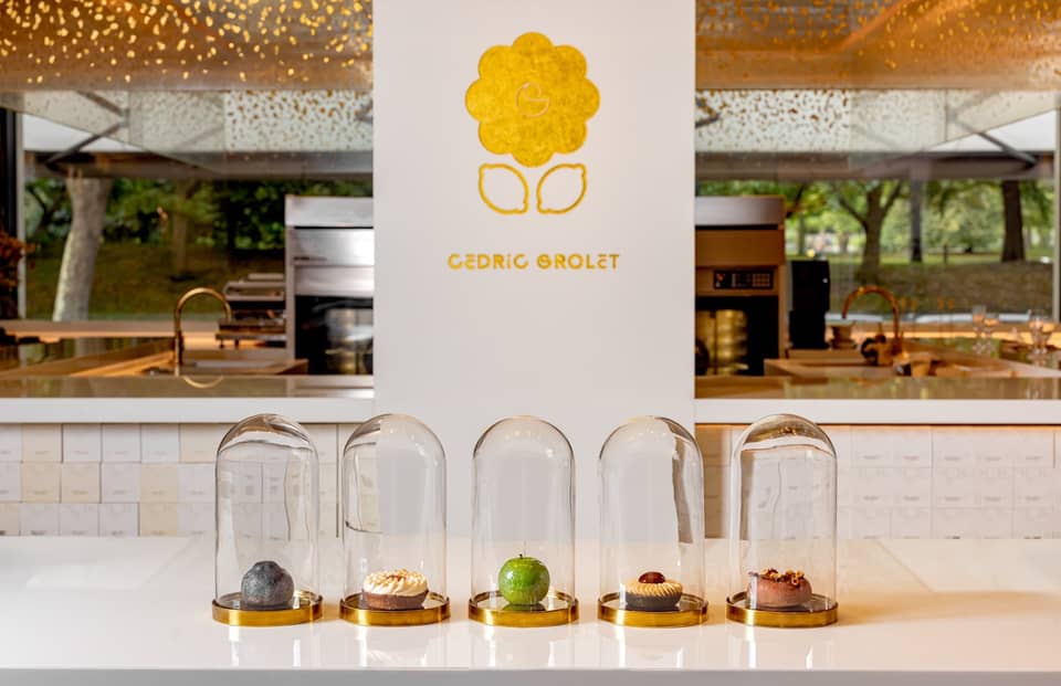 Five exquisite pastries encased in glass domes, elegantly displayed on a minimalist white counter with Cedric Grolet's golden logo in the background.