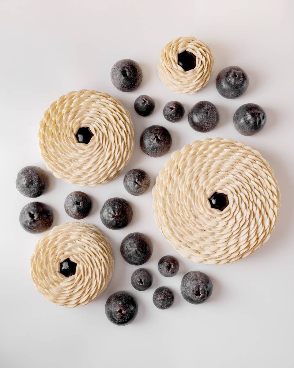 An artistic arrangement of intricately layered pastries with spiral patterns, paired with dark, round fruit-like elements on a pristine white background, creating a visually striking contrast.