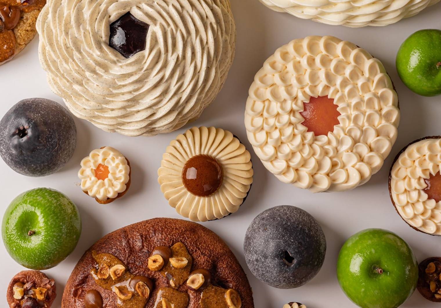A vibrant assortment of pastries and fruit-like confections arranged on a white background, featuring spiraled cream textures, glossy green and dark accents, and nut-studded elements in a harmonious display.