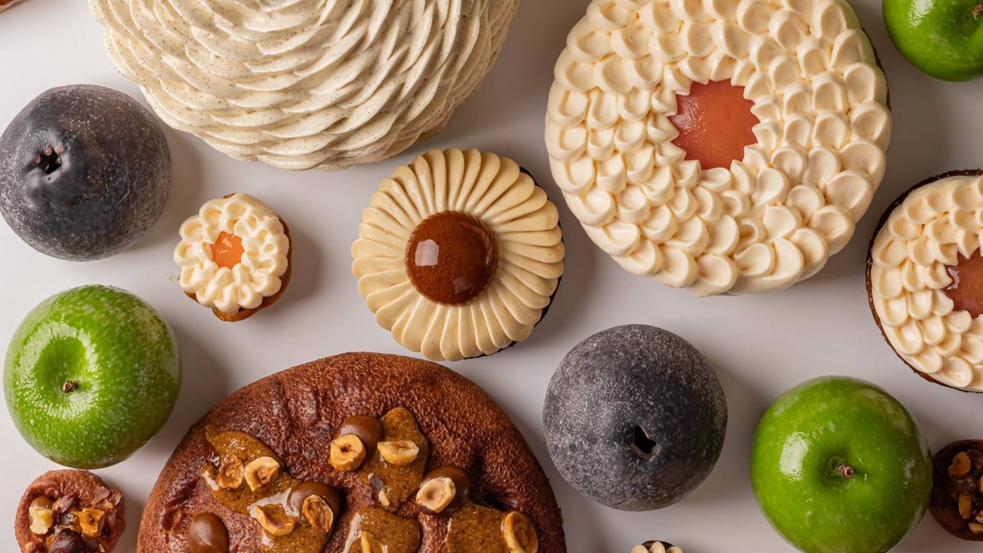 A vibrant assortment of pastries and fruit-like confections arranged on a white background, featuring spiraled cream textures, glossy green and dark accents, and nut-studded elements in a harmonious display.
