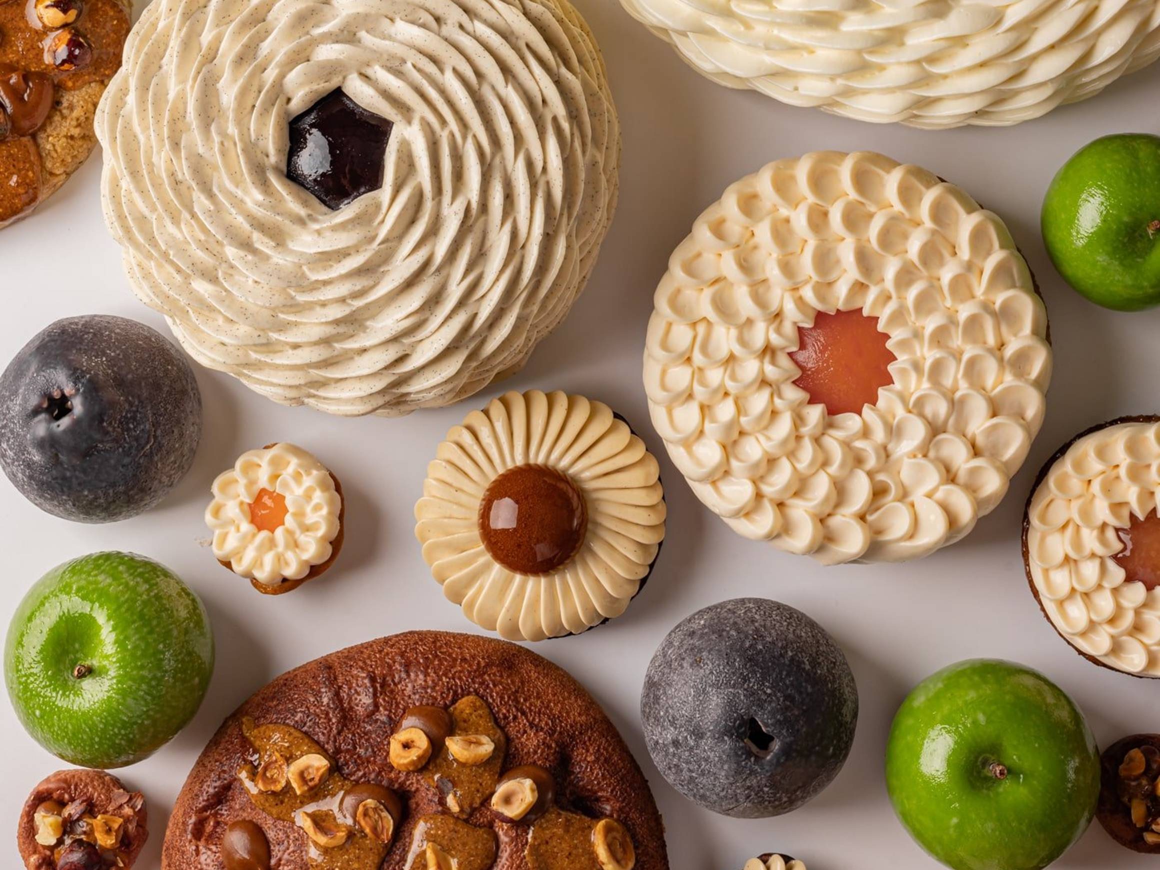 A vibrant assortment of pastries and fruit-like confections arranged on a white background, featuring spiraled cream textures, glossy green and dark accents, and nut-studded elements in a harmonious display.