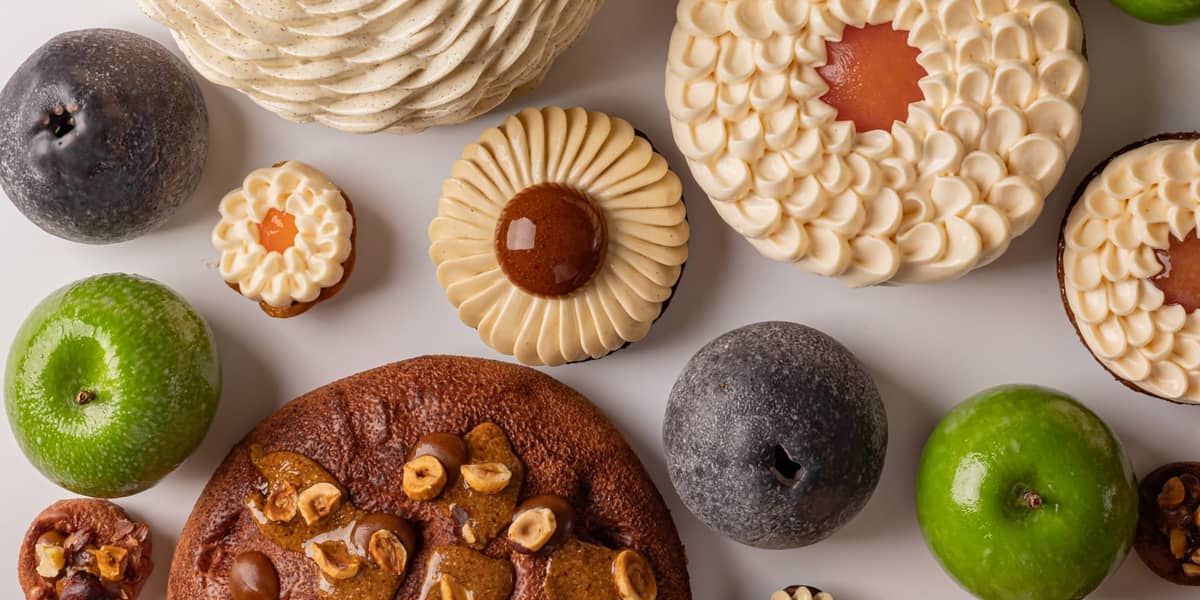 A vibrant assortment of pastries and fruit-like confections arranged on a white background, featuring spiraled cream textures, glossy green and dark accents, and nut-studded elements in a harmonious display.