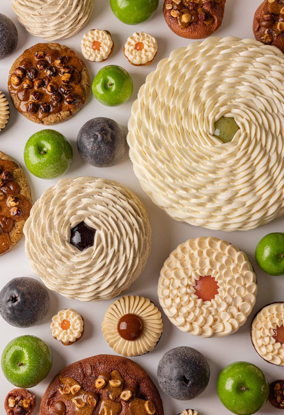 A vibrant assortment of pastries and fruit-like confections arranged on a white background, featuring spiraled cream textures, glossy green and dark accents, and nut-studded elements in a harmonious display.