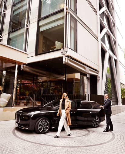 A woman wearing a beige coat and white trousers is walking past a black car. A man is closing the door of the car.