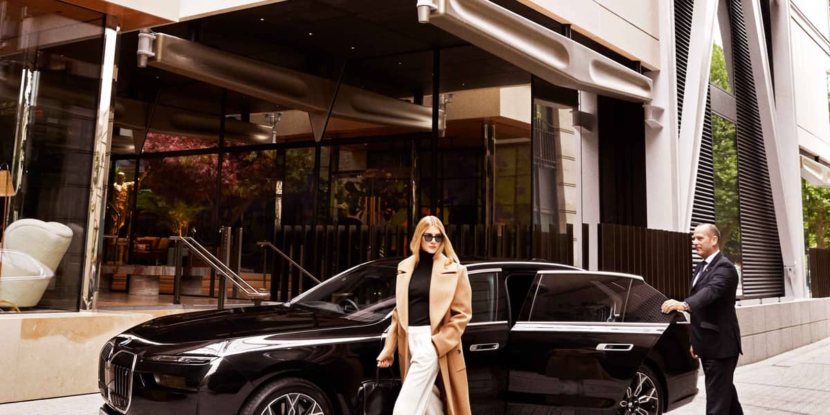 A woman wearing a beige coat and white trousers is walking past a black car. A man is closing the door of the car.