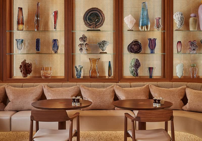 Two round wooden tables and two wooden chairs are in the centre. A couch with pink cushions is in the background. Behind the couch are a number of coloured ornaments behind a glass cupboard.