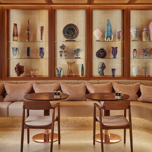 Two round wooden tables and two wooden chairs are in the centre. A couch with pink cushions is in the background. Behind the couch are a number of coloured ornaments behind a glass cupboard.