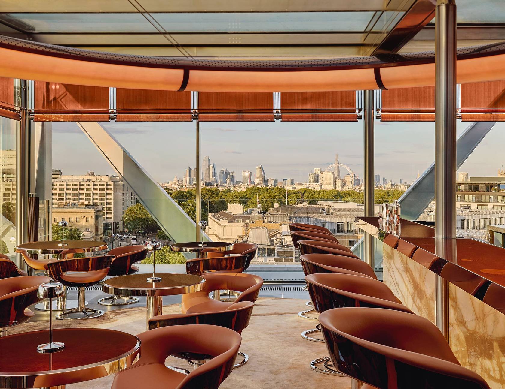 The Emory Rooftop bar, circular tables and brown leather chairs are placed in a bright room with views of the London skyline